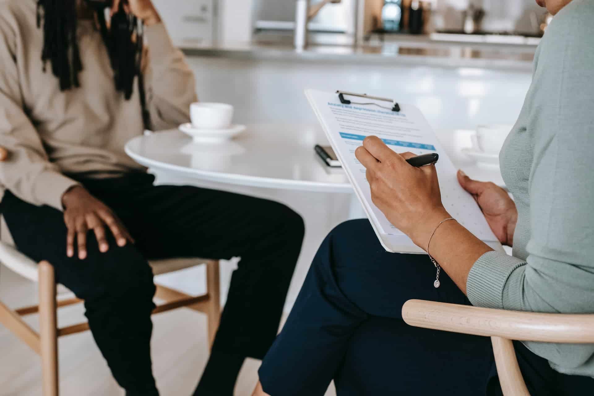 Two people sitting in chairs, faces cropped out of photo, one with a clipboard giving a performance review of the other