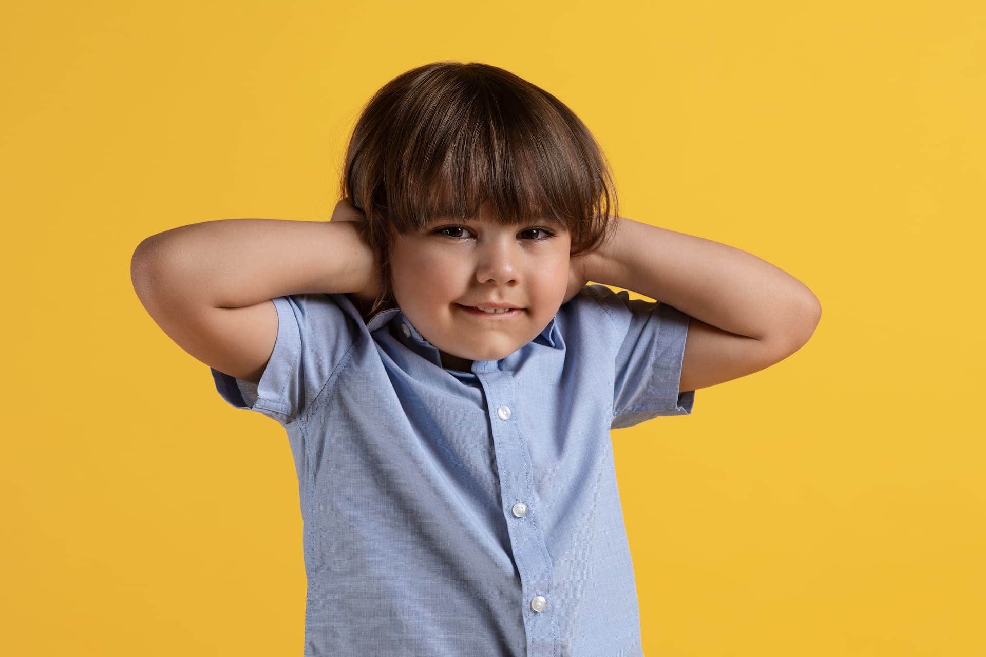 Close up portrait of annoyed little boy covering ears with hands and looking at camera, orange studio background.