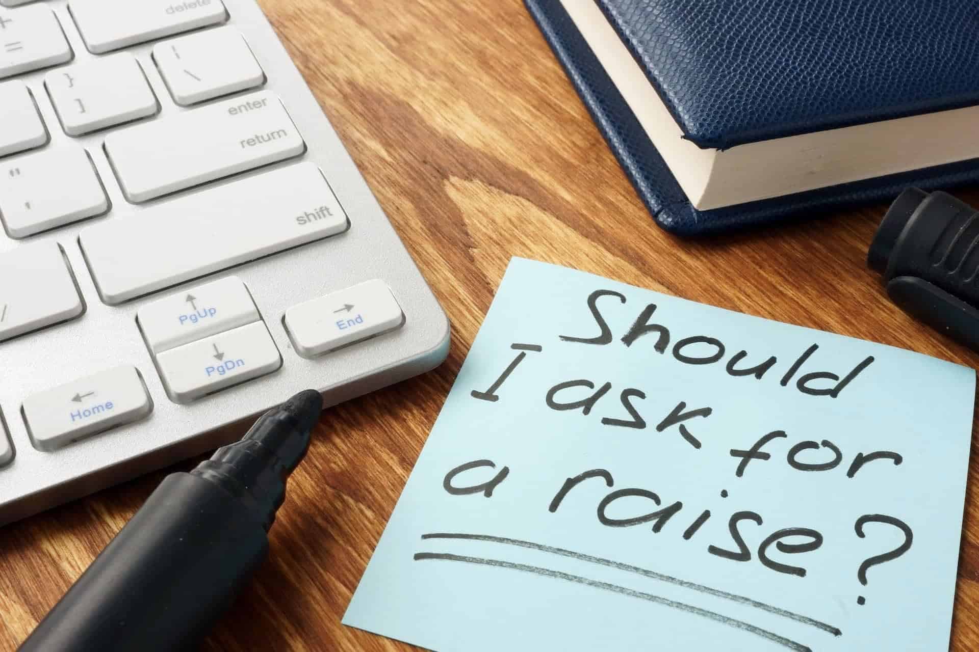 How to ask for a raise: Office desk with office items scattered and a post it note that reads, Should I ask for a raise?